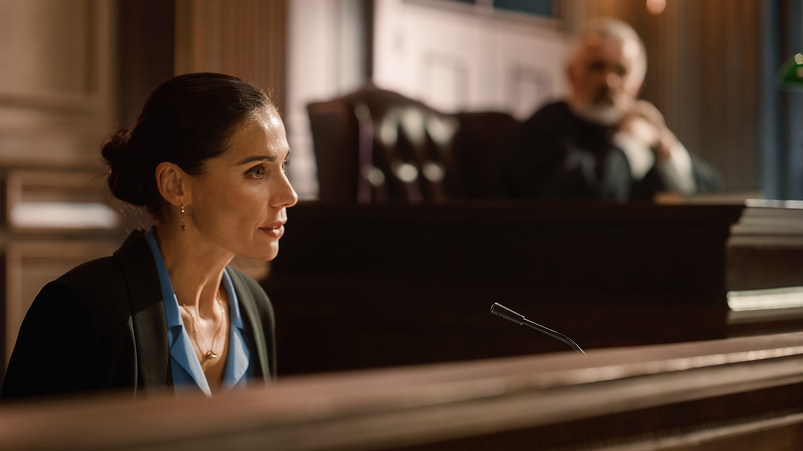 Female expert witness testifying in a court proceeding with the judge in the background listening to the testimony.