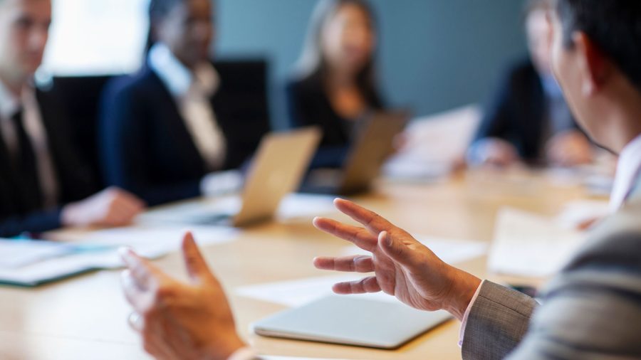 A group of business professionals sit around a conference table, engaged in discussion. Laptops, papers, and pens are visible on the table, and one person gestures while speaking. The background is softly blurred.