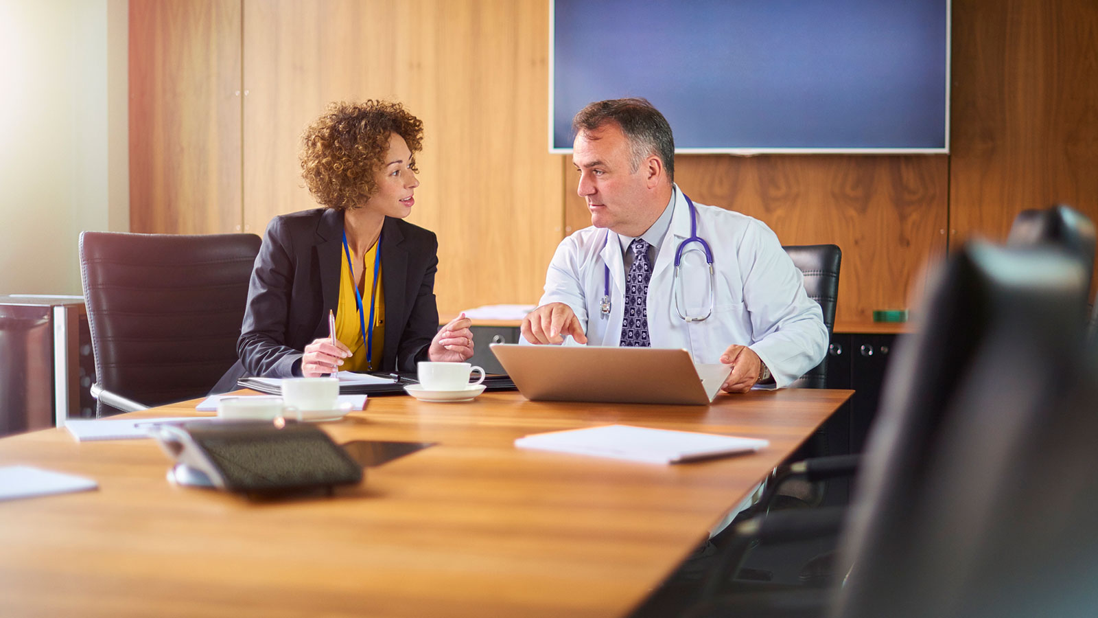 A doctor with a stethoscope and a woman in business attire sit at a conference table, discussing something on a laptop. Papers, coffee cups, and office supplies are on the table in a modern meeting room.