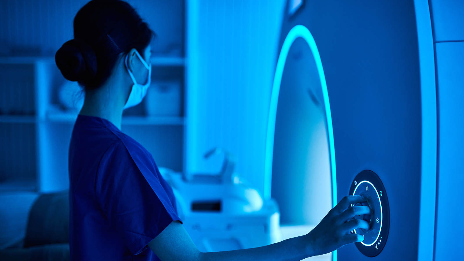 A healthcare worker in scrubs and a mask operates an MRI machine, pressing a button on the control panel in a blue-lit medical room.