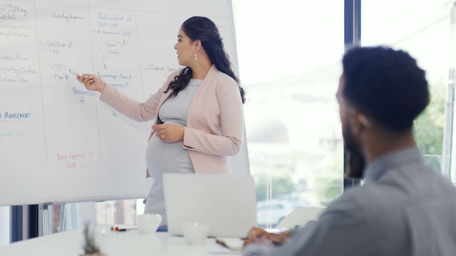 A pregnant woman stands by a whiteboard, pointing at notes while presenting to a seated colleague in a modern office setting.