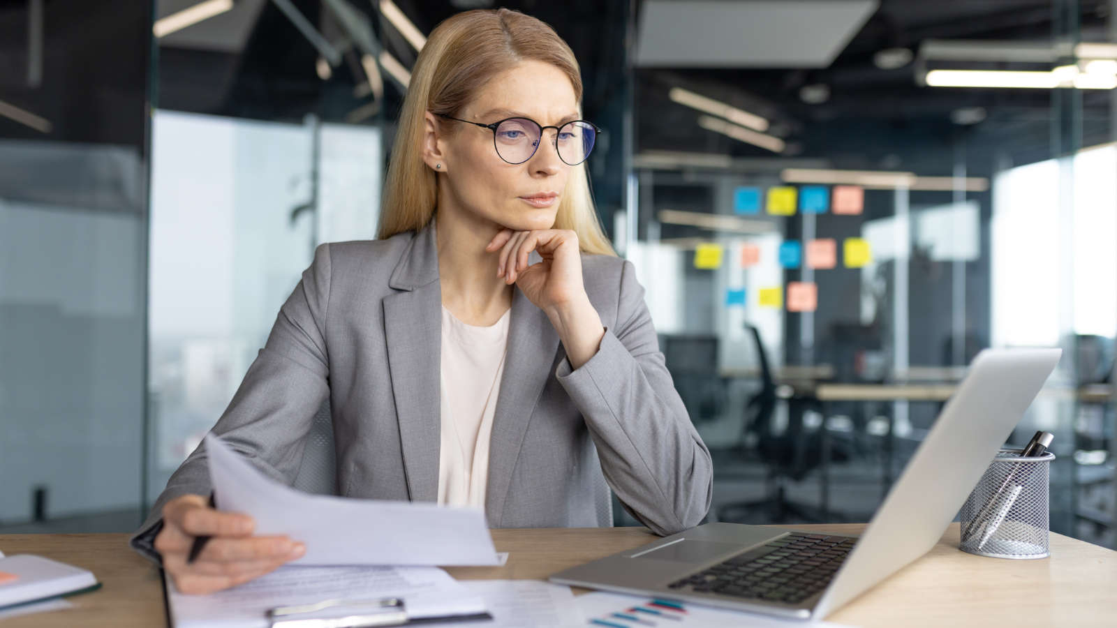 A woman in glasses and a gray blazer sits at a desk in a modern office, looking thoughtfully at a laptop while holding papers. Colorful sticky notes are visible on the glass wall behind her.