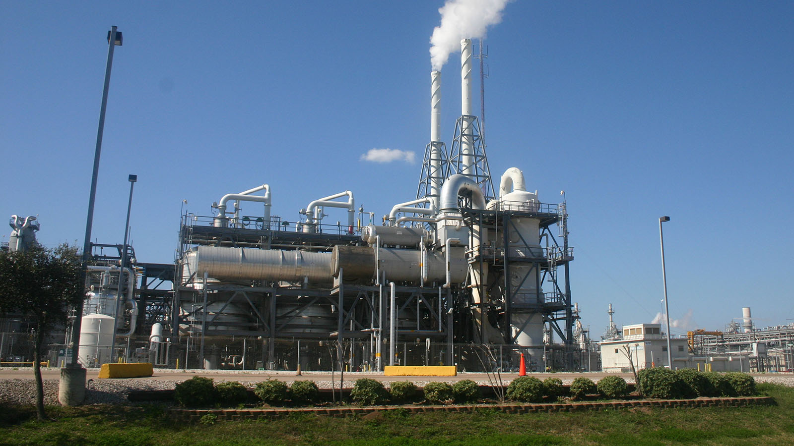 Industrial chemical plant with tall smokestacks emitting white smoke against a clear blue sky.