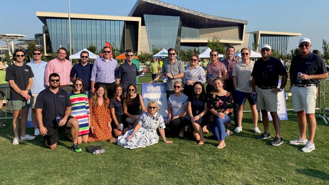 A large group of adults poses for a photo on a grassy lawn in front of a modern glass building, with a sponsor sign for McAfee displayed in the center.